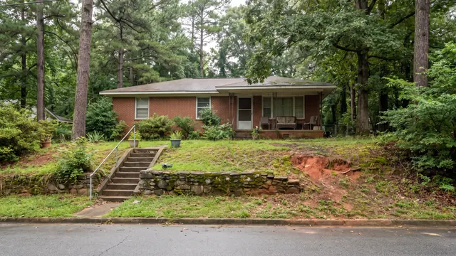 A brick ranch house set above the road on a sloped wooded lot with steps up from the pavement and exposed red clay, typical of everyday residential Birmingham, Alabama