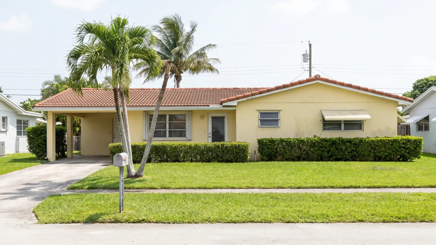 A single storey concrete block ranch house with a barrel tile roof, carport and palm trees, typical of everyday residential Broward County, Florida