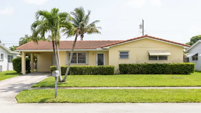 A single storey concrete block ranch house with a barrel tile roof, carport and palm trees, typical of everyday residential Broward County, Florida