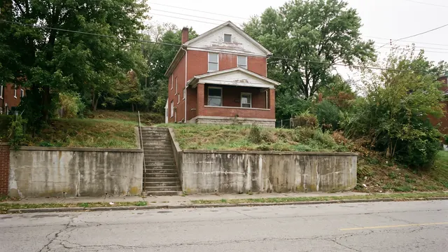 A narrow two storey brick house standing above the street on a hillside lot, with a concrete retaining wall and steps climbing to the front door, typical of everyday residential Cincinnati, Ohio