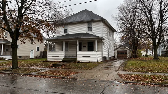 A white American Foursquare house with a full width front porch and a detached garage, bare autumn trees and a wet street, typical of everyday residential Cleveland, Ohio