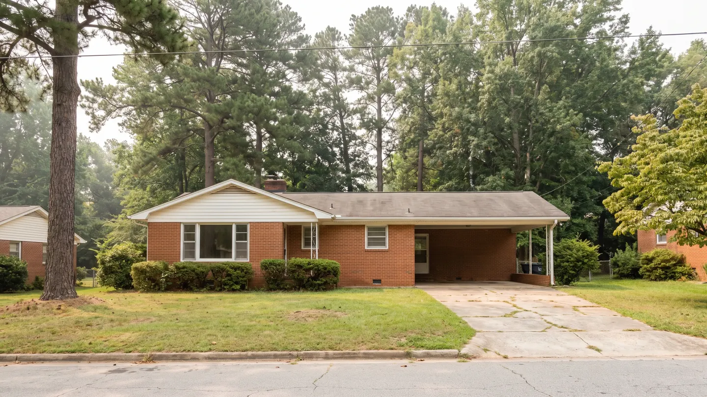 A red brick ranch house with a carport and a picture window, backed by tall loblolly pines, typical of everyday residential Greensboro, North Carolina
