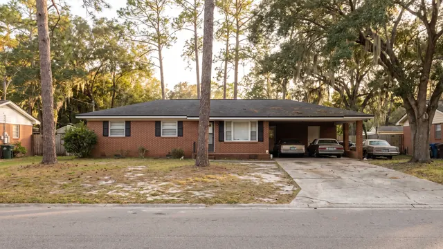 A red brick ranch house with a carport, set among tall pines and live oaks draped in Spanish moss, typical of everyday residential Jacksonville, Florida