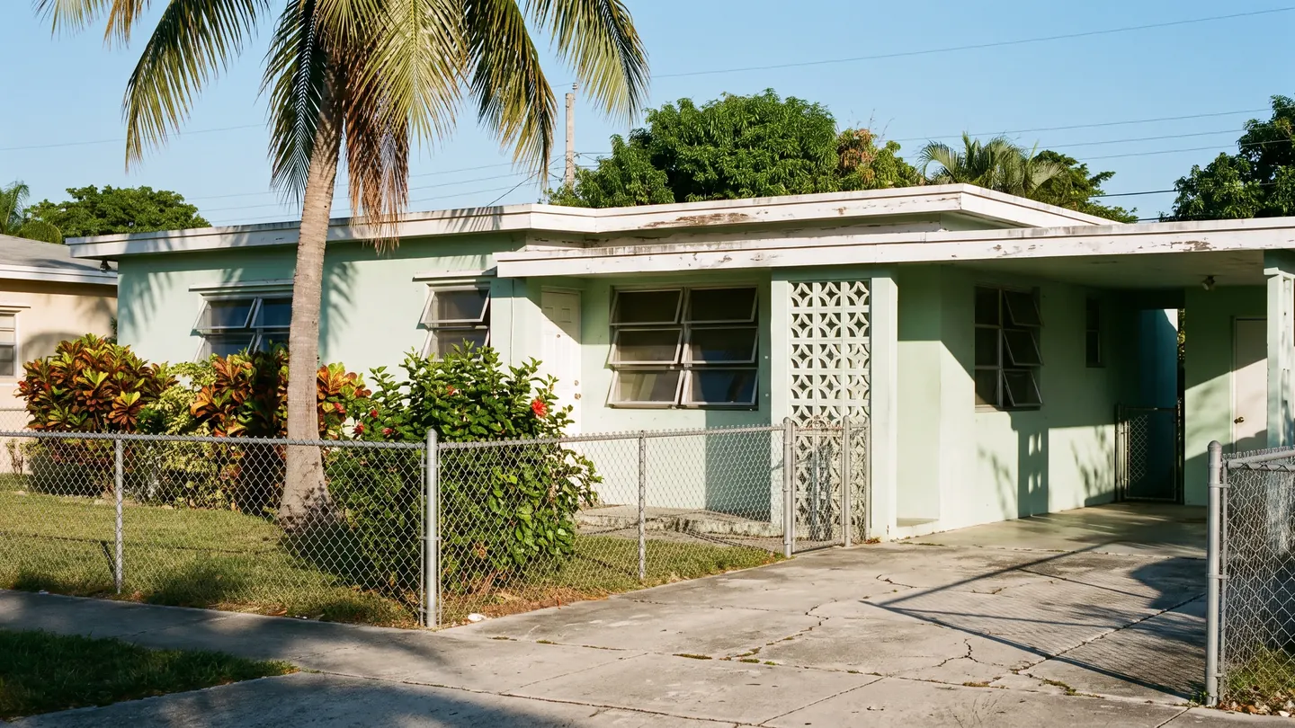 A mid century mint green stucco house with jalousie windows, a decorative breeze block screen and a chain link fence, typical of everyday residential Miami-Dade County, Florida