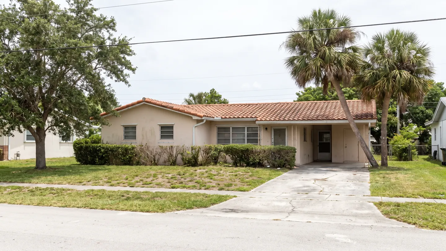 A single storey stucco ranch house with a weathered barrel tile roof, a carport, a cracked concrete driveway, a clipped hedge and sabal palms on a patchy lawn, on an ordinary flat residential street in Palm Beach County, Florida