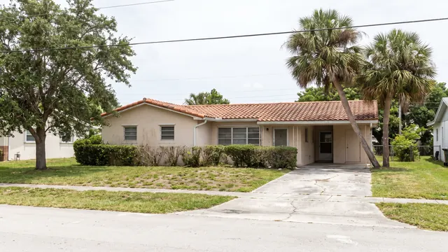 A single storey stucco ranch house with a weathered barrel tile roof, a carport, a cracked concrete driveway, a clipped hedge and sabal palms on a patchy lawn, on an ordinary flat residential street in Palm Beach County, Florida