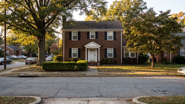 A two storey red brick colonial style house with black shutters and a low hedge on an established tree lined street, typical of everyday residential Winston-Salem, North Carolina